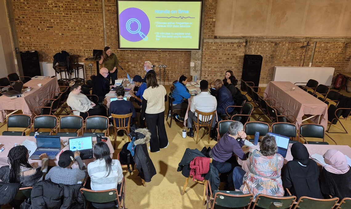 Birds eye view of a room of people from charities and community organisations. There is a data presentation showing on a large screen at the front of the room. Some people are chatting, others have their laptops on tables in front of them